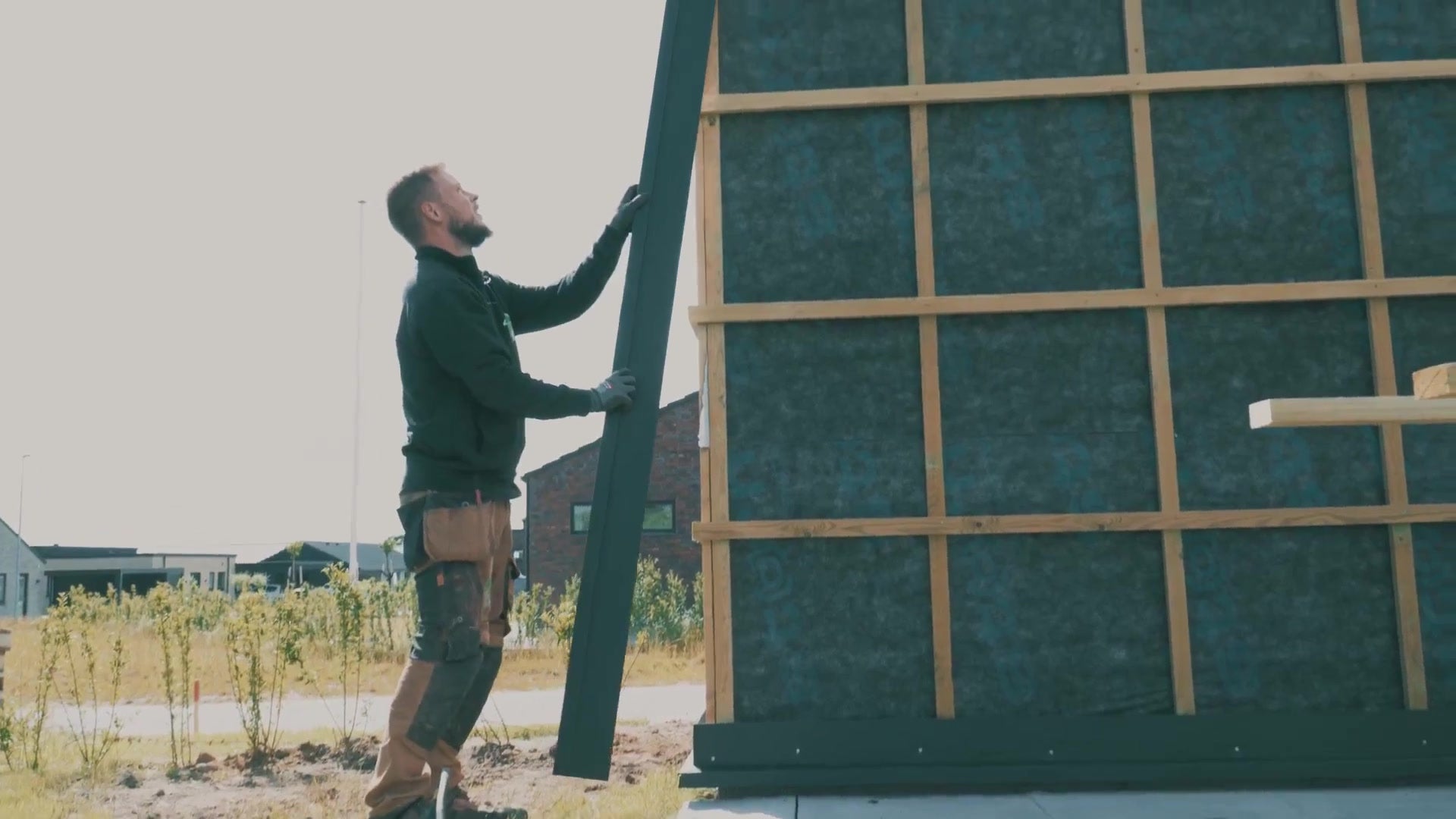 Carpenter lifting a black corner profile into place against a membrane-covered garden shed wall